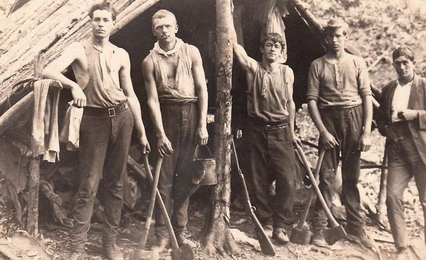 five young loggers standing in front of a handmade wooden shelter holding axes