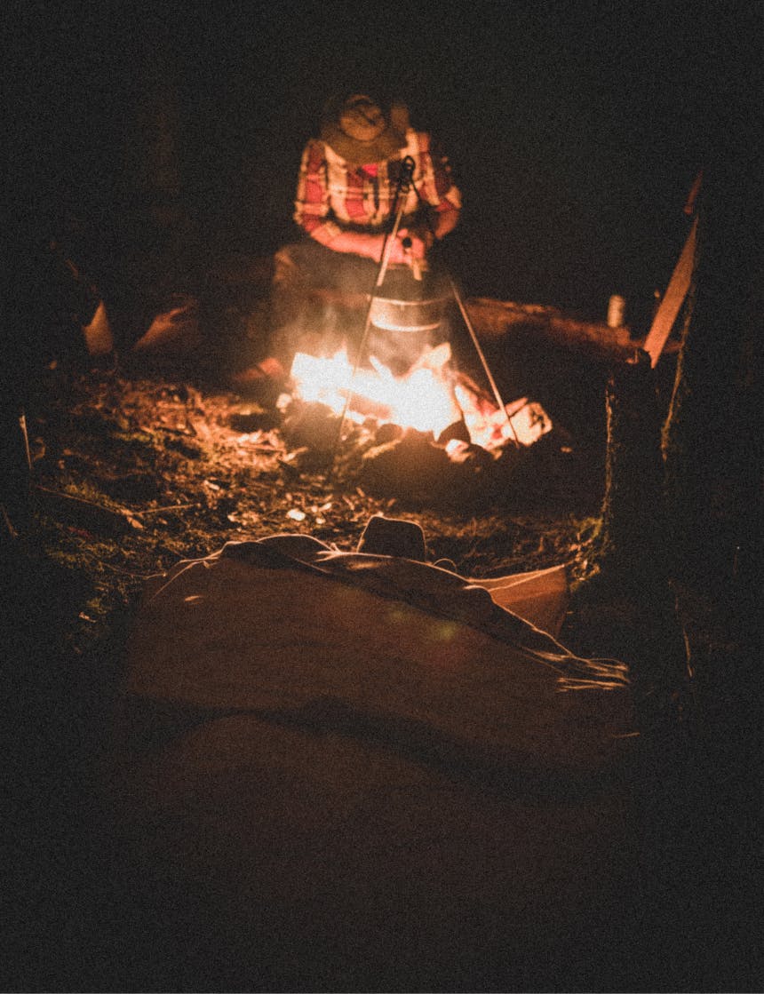 man in red and white plaid shirt sitting by a fire with a pot hanging over the flames