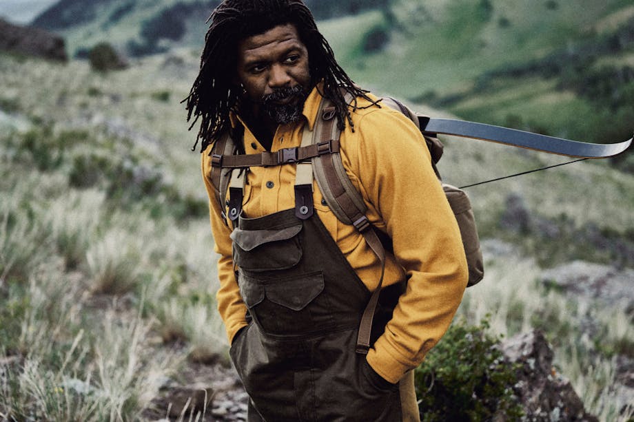 ray livingston in mustard shirt and filson backpack with crossbow on back standing in a mountain meadow