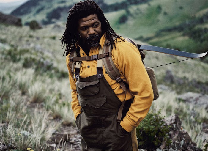 ray livingston in mustard shirt and filson backpack with crossbow on back standing in a mountain meadow