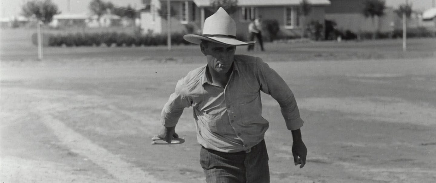 man wearing a white cowboy hat and button up shirt throwing horseshoes in a dirt field