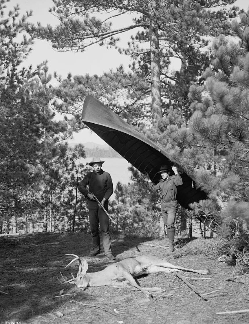 one man holding a canoe over his head next to a man holding a rifle standing over a downed deer