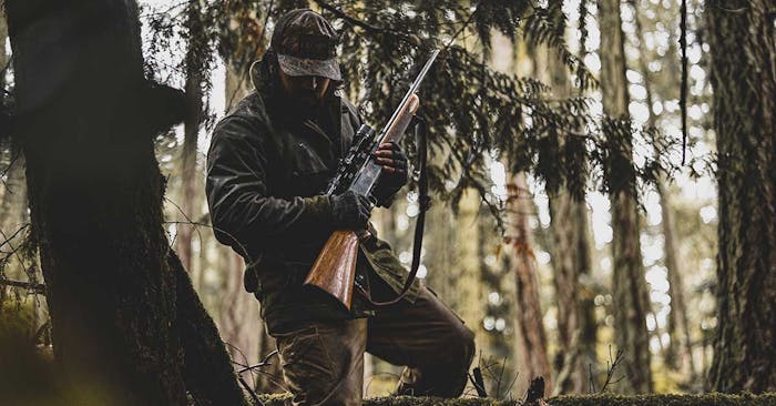 person in green coat and hat holding a rifle walking through a pine tree forest