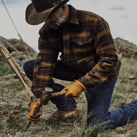 man in leather hat and filson denim secures a stake into grassy ground