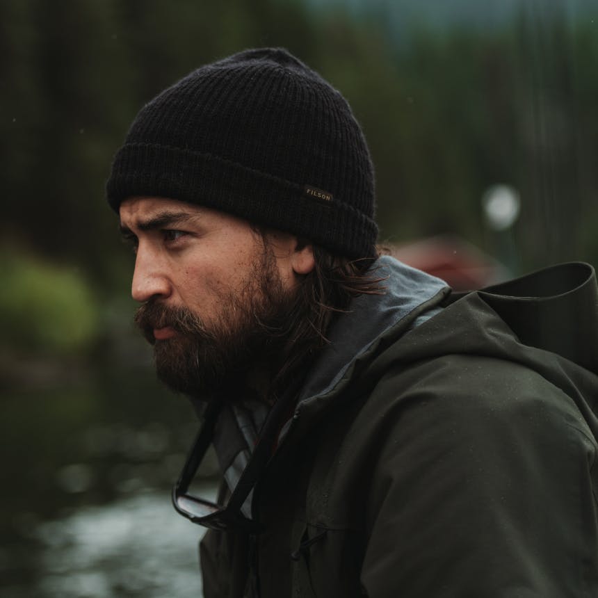 Man in black filson beanie and matte green jacket in forest with river in background