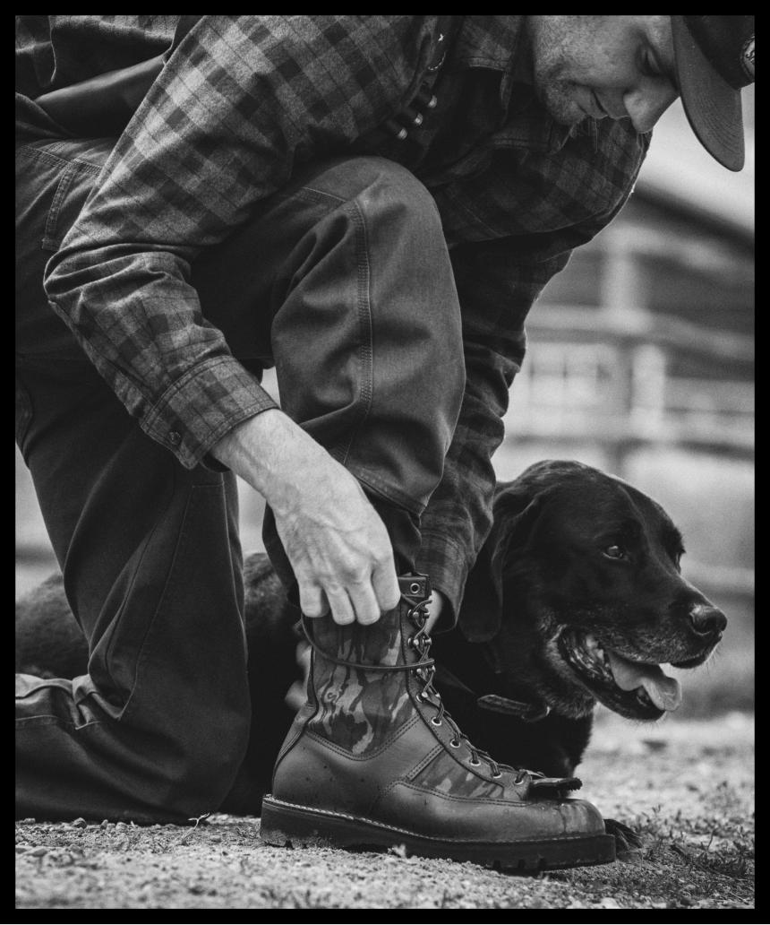 man kneels to tie boot in plaid shirt next to black dog