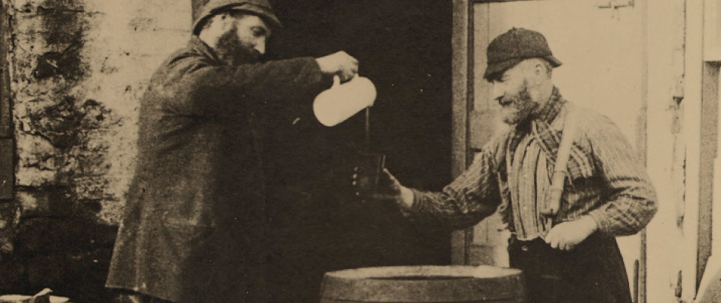 black and white image of man in black coat pours a drink out of a white pitcher for man in suspenders and plaid shirt