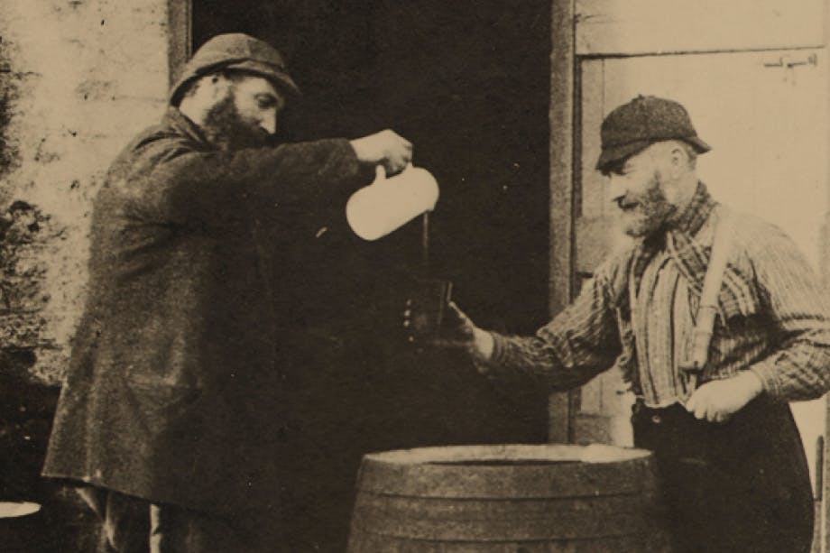 black and white image of man in black coat pours a drink out of a white pitcher for man in suspenders and plaid shirt