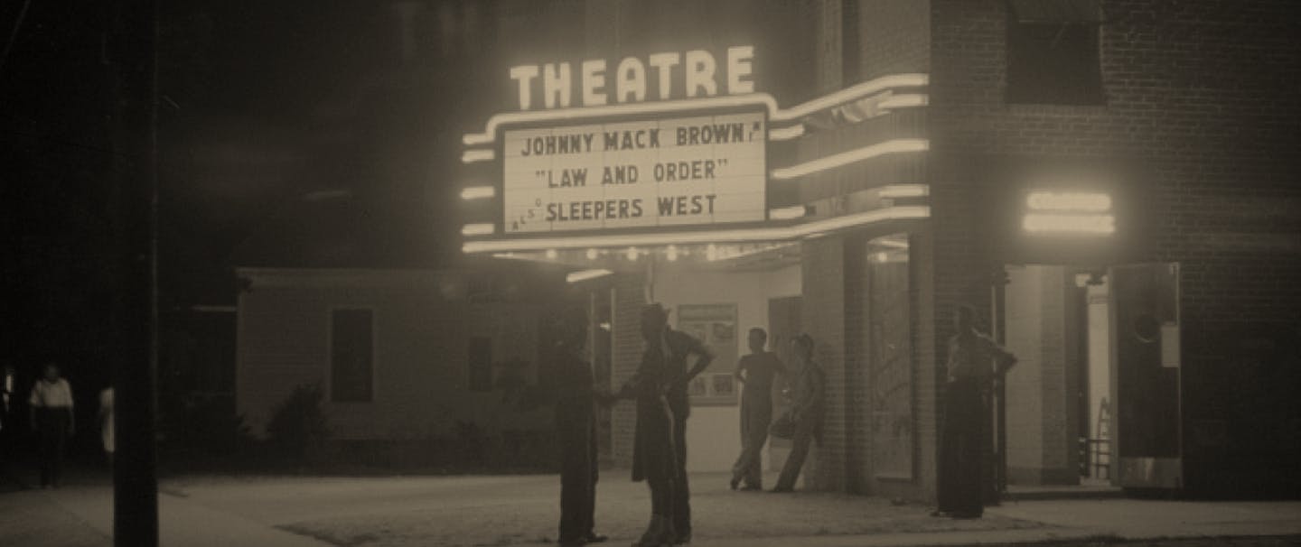 Black and white image of people standing in front of theatre with marquee reading 