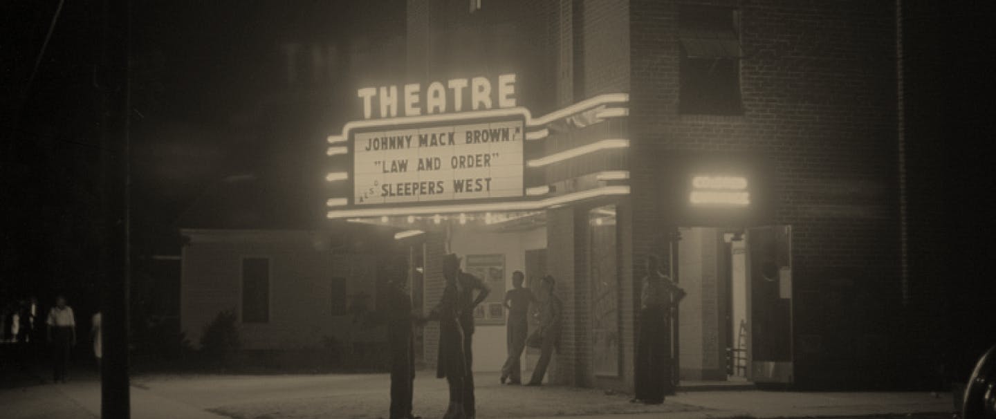 Black and white image of people standing in front of theatre with marquee reading 