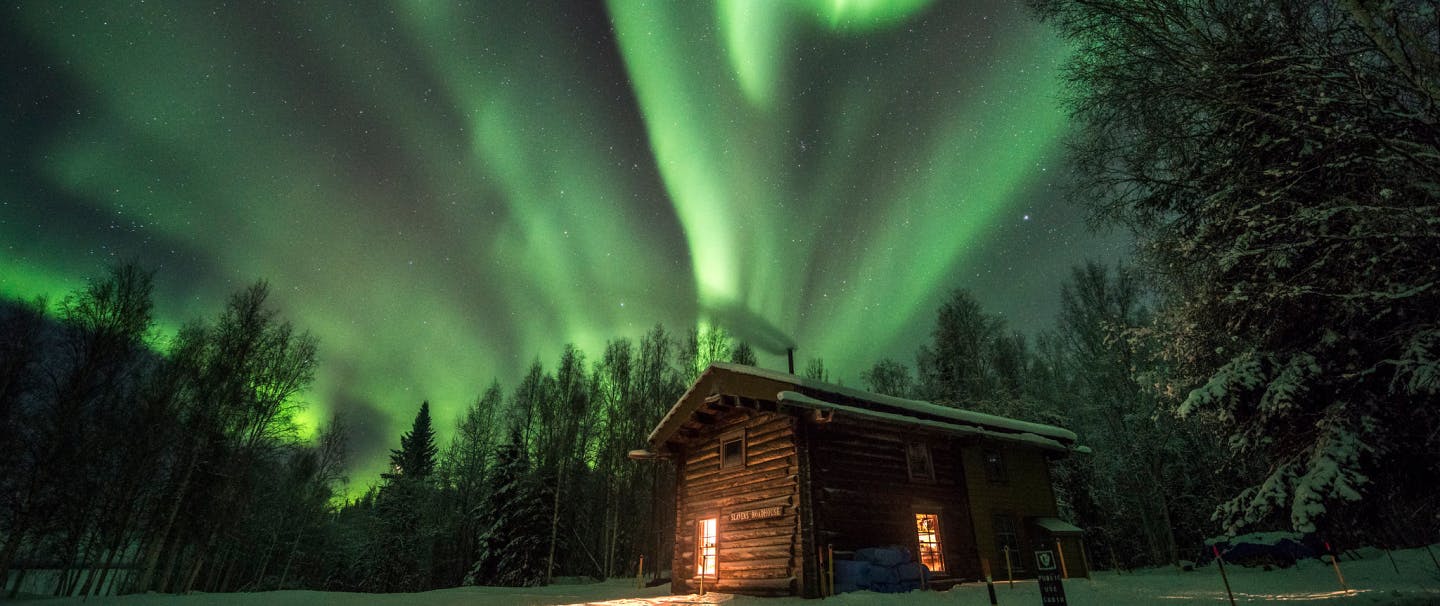 snow covered lit cabin in forest with green northern lights above