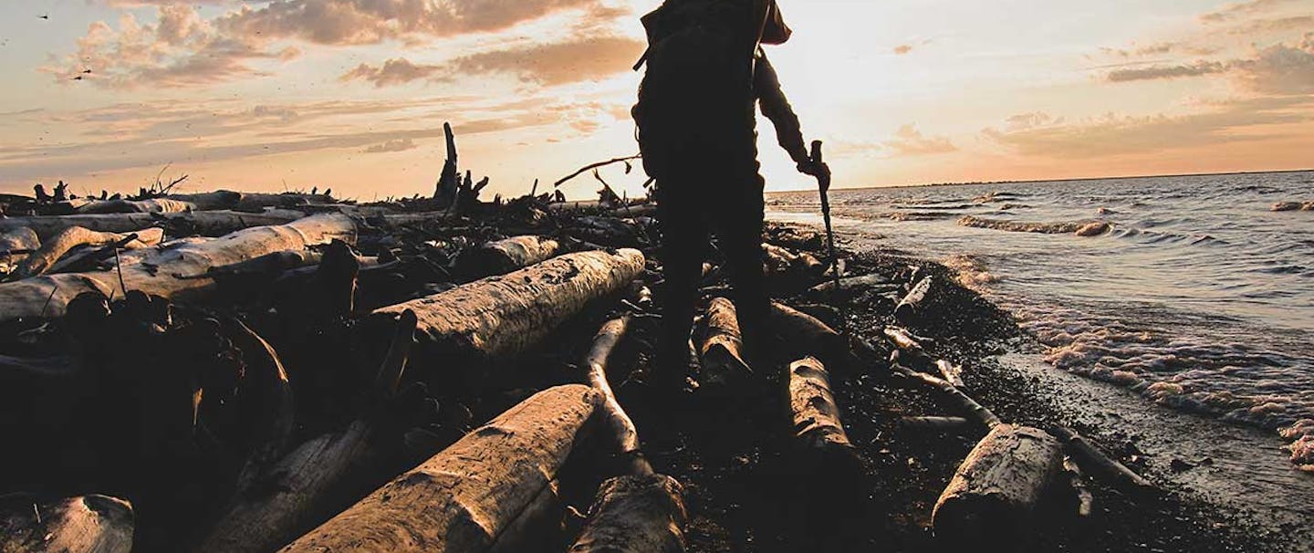 Silhouette of man with walking pole and backpacking backpack walking along beach with downed logs