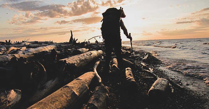 Silhouette of man with walking pole and backpacking backpack walking along beach with downed logs