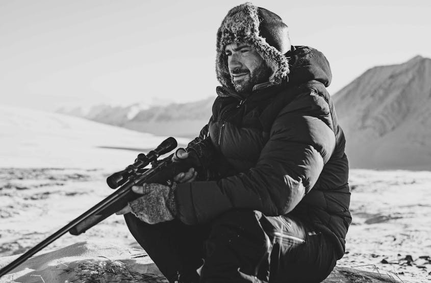 man in fur lined ushanka hat and black parka sitting in a snowy arctic plain holding a rifle with mountains in the background