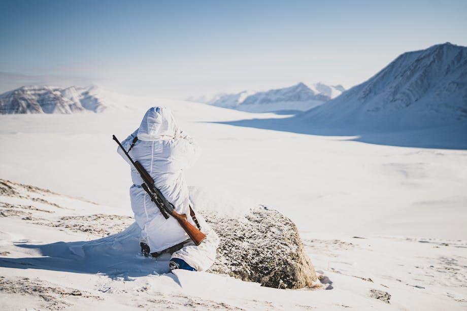 person in white parka looking out over a vast snowy plain with mountains with rifle over his back