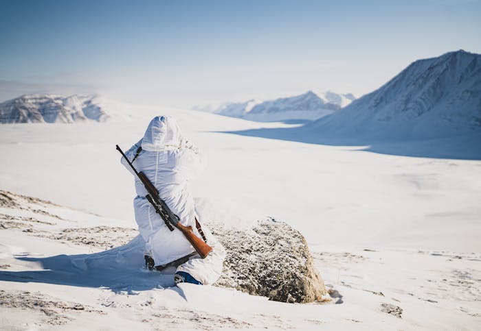 person in white parka looking out over a vast snowy plain with mountains with rifle over his back