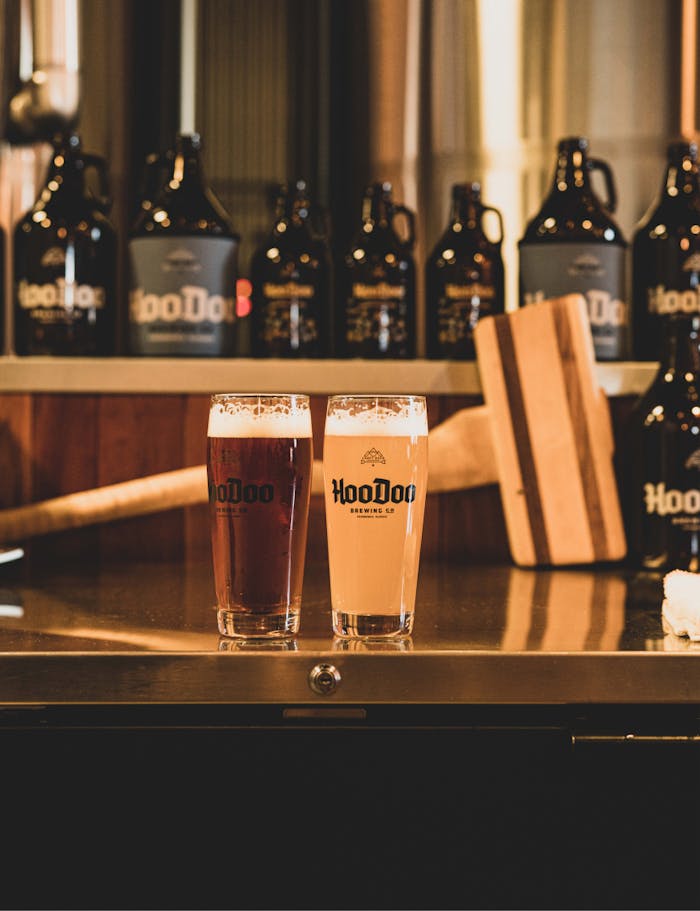 two pint glasses of hoodoo beer on a stainless steel counter with growlers of different sizes behind them