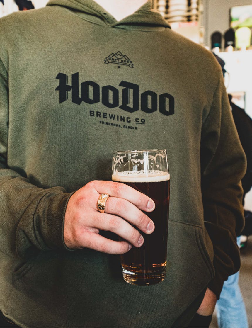 person with a gold ring on their finger holding a dark beer wearing a green hoodoo hoodie