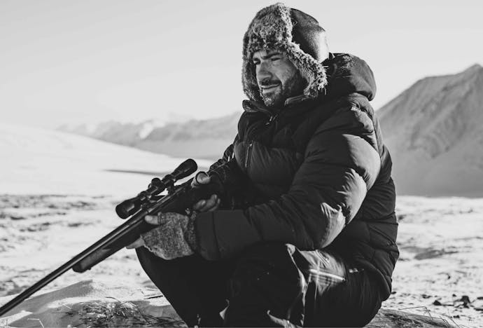 person in a fur hat with ear flaps, black parka, holding a rifle sitting in a snowy field with mountains in the background