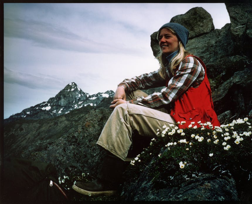 woman in red vest, white pants, and orange and white plaid shirt sitting on a rocky mountain escarpment next to some small white flowers