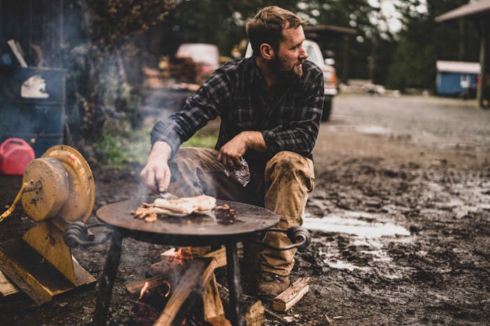 person tending to fish cooking on a metal table over a wood fire in a muddy plot