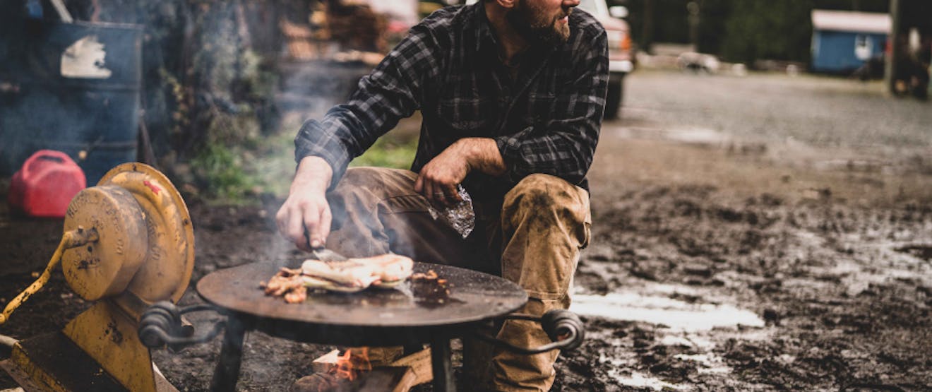 person tending to fish cooking on a metal table over a wood fire in a muddy plot
