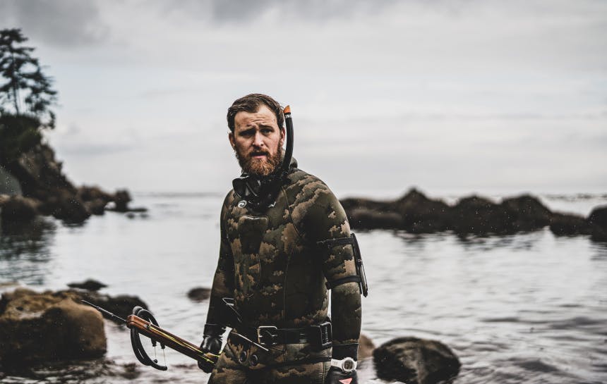 man in wetsuit and snorkeling gear exiting the ocean holding a harpoon