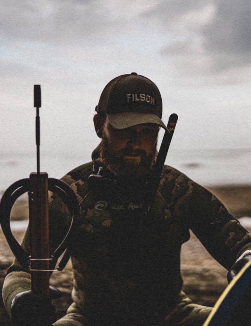 person in brown filson hat sitting on beach in wetsuit holding a harpoon gun