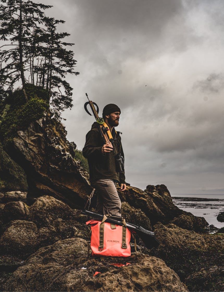 man in black beanie standing on beach holding a harpoon gun over his shoulder next to an orange dry filson bag