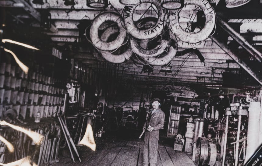 black and white image of man standing in a workshop under a collection of life floats hanging by the rafters