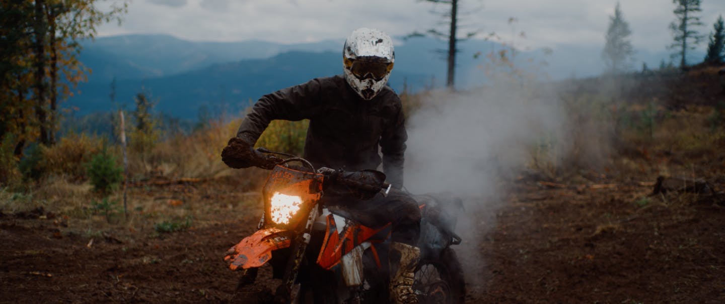 person in mud spattered white helmet riding a dirt bike in a pine forest clearing kicking up smoke from exhaust