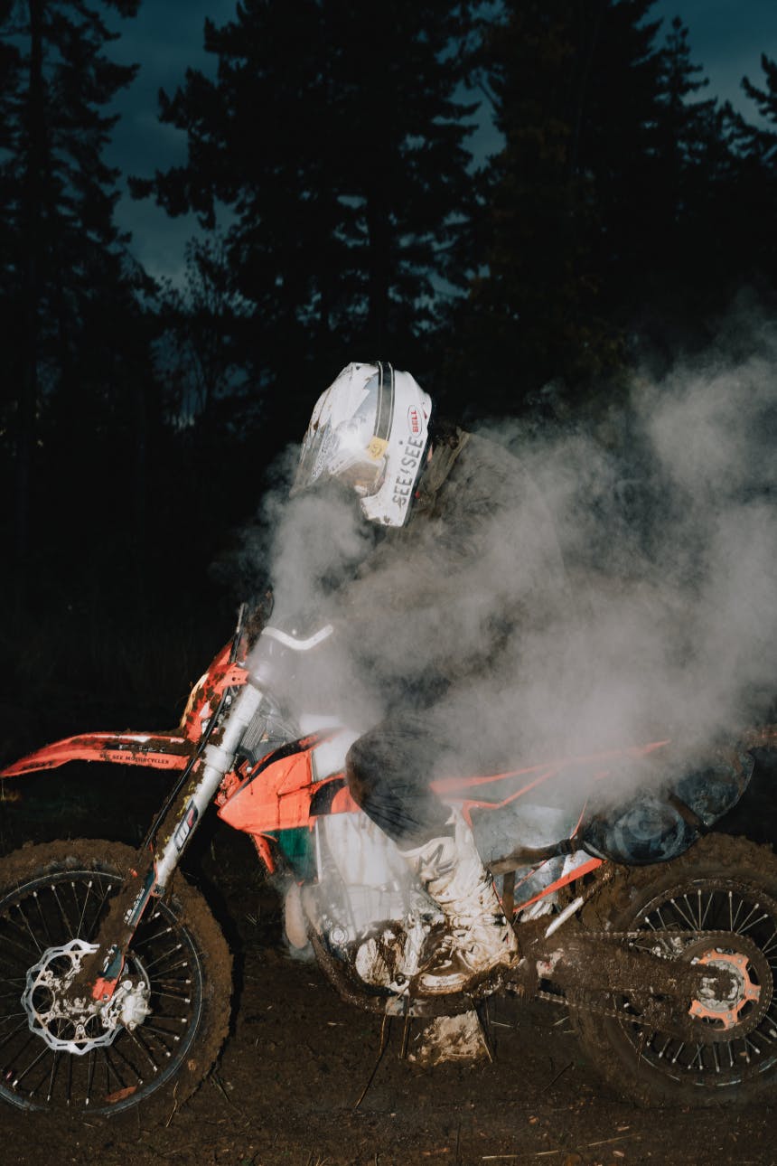 person in white motorcycle helmet sitting on a smoking orange motorcycle covered in mud