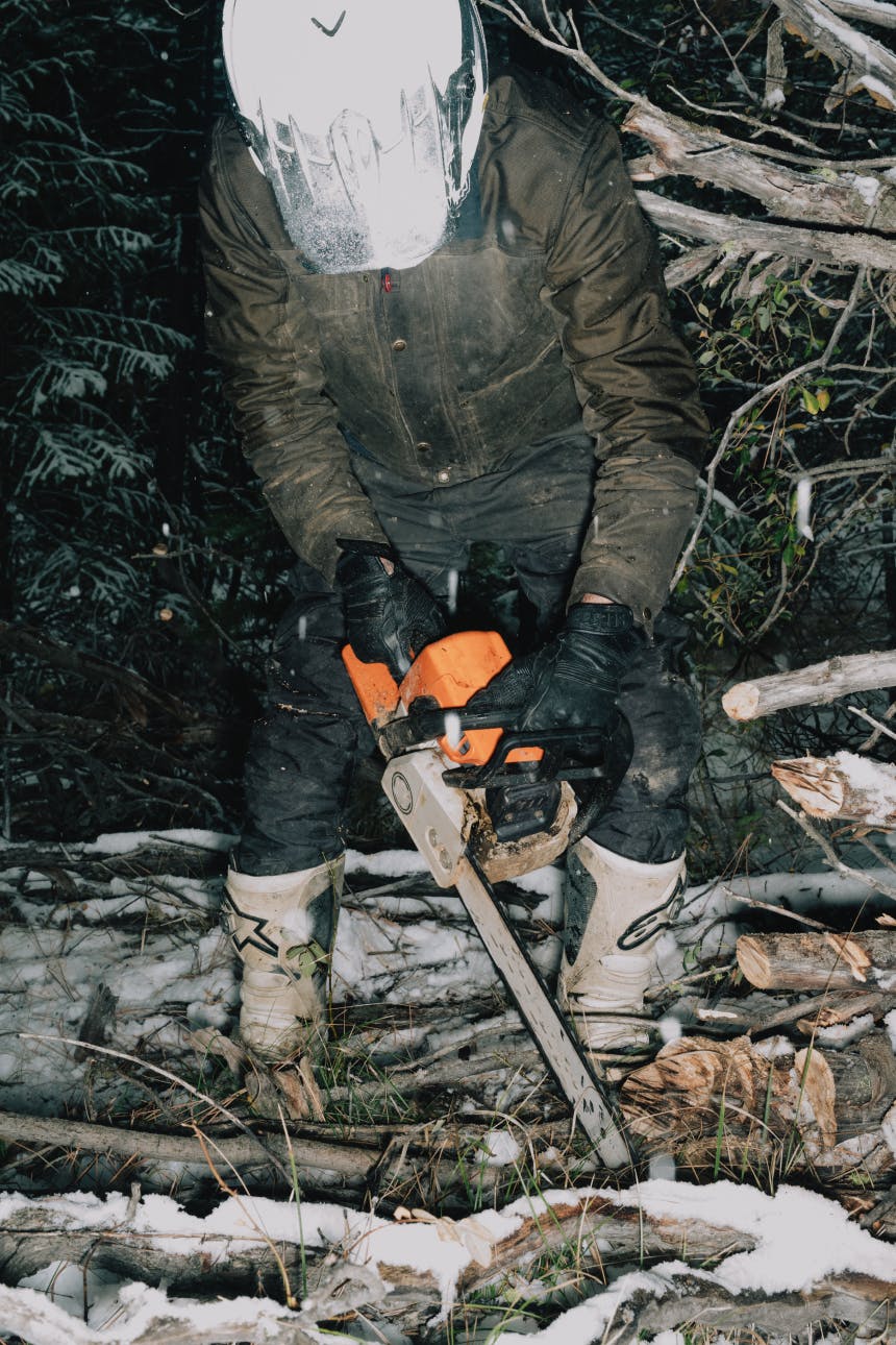 person in brown coat and black pants and motorcycle helmet cutting wood with an orange chainsaw