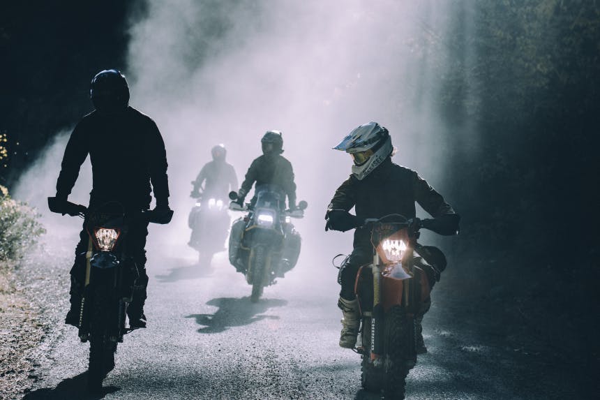 four people riding their motorcycles down a mud road in the forest bathed in fog