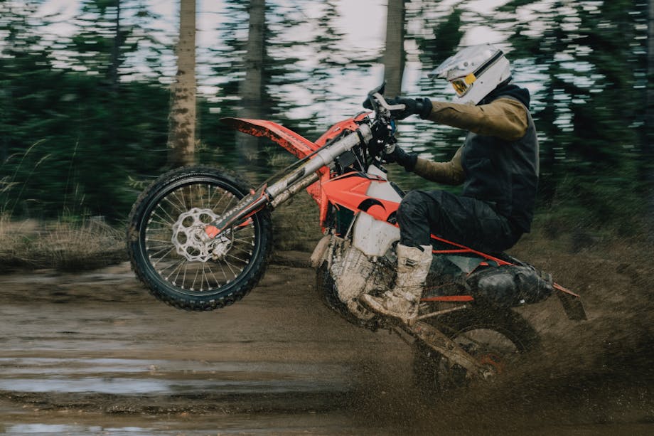 person popping a wheelie on an orange motorcycle on a muddy road in the forest