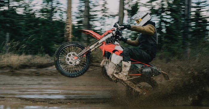 person popping a wheelie on an orange motorcycle on a muddy road in the forest