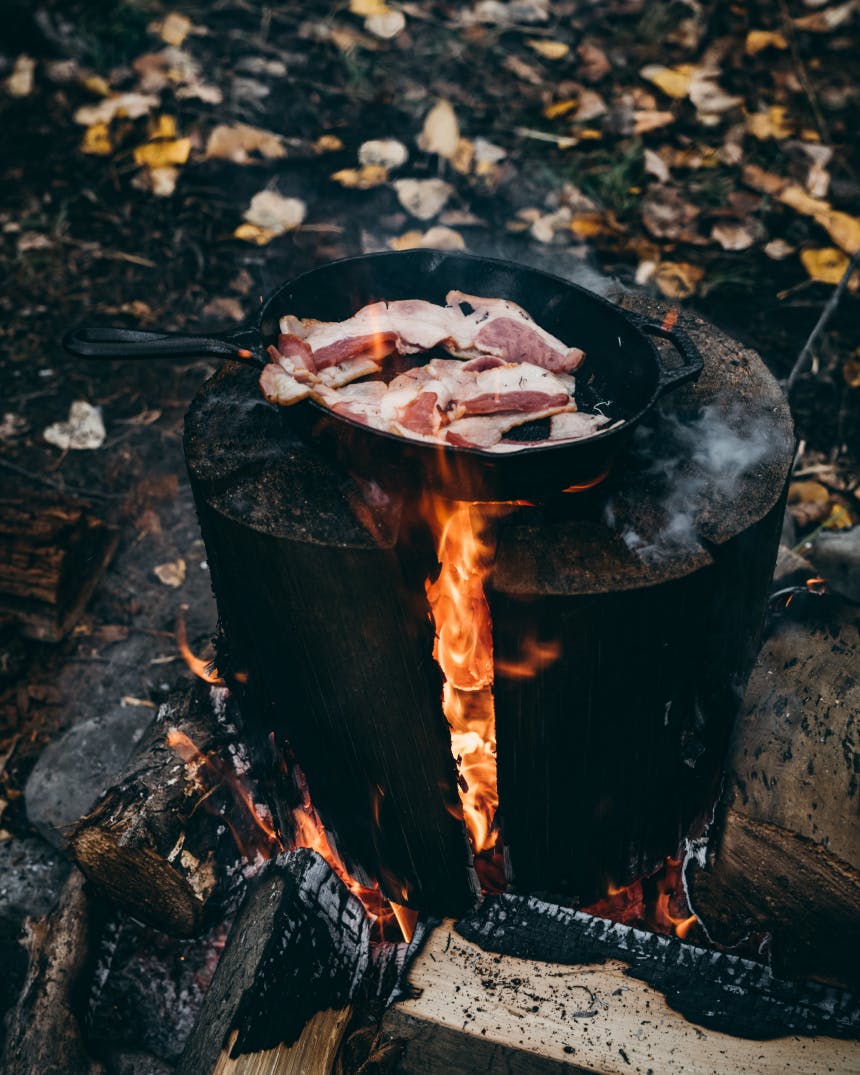 cuts of meat cooking in a cast iron skillet on top of a rocket stove made out of a standing log