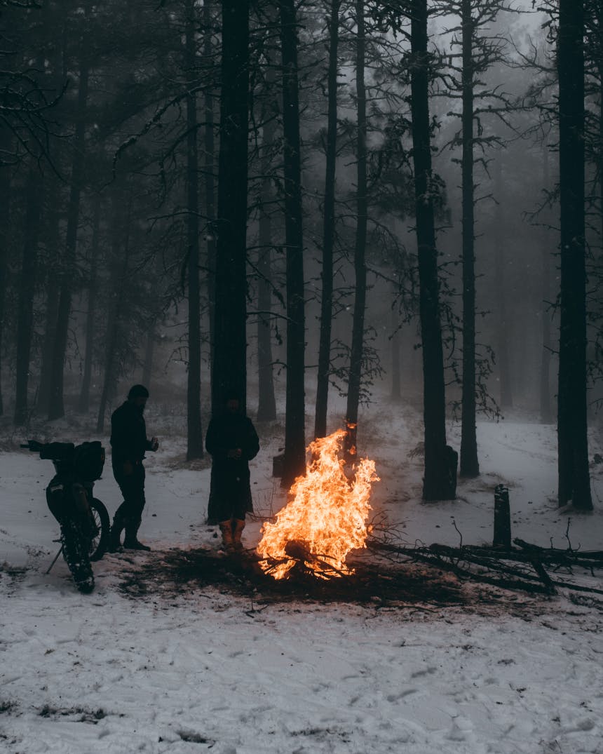 people in a snowy campsite stand next to a very large fire