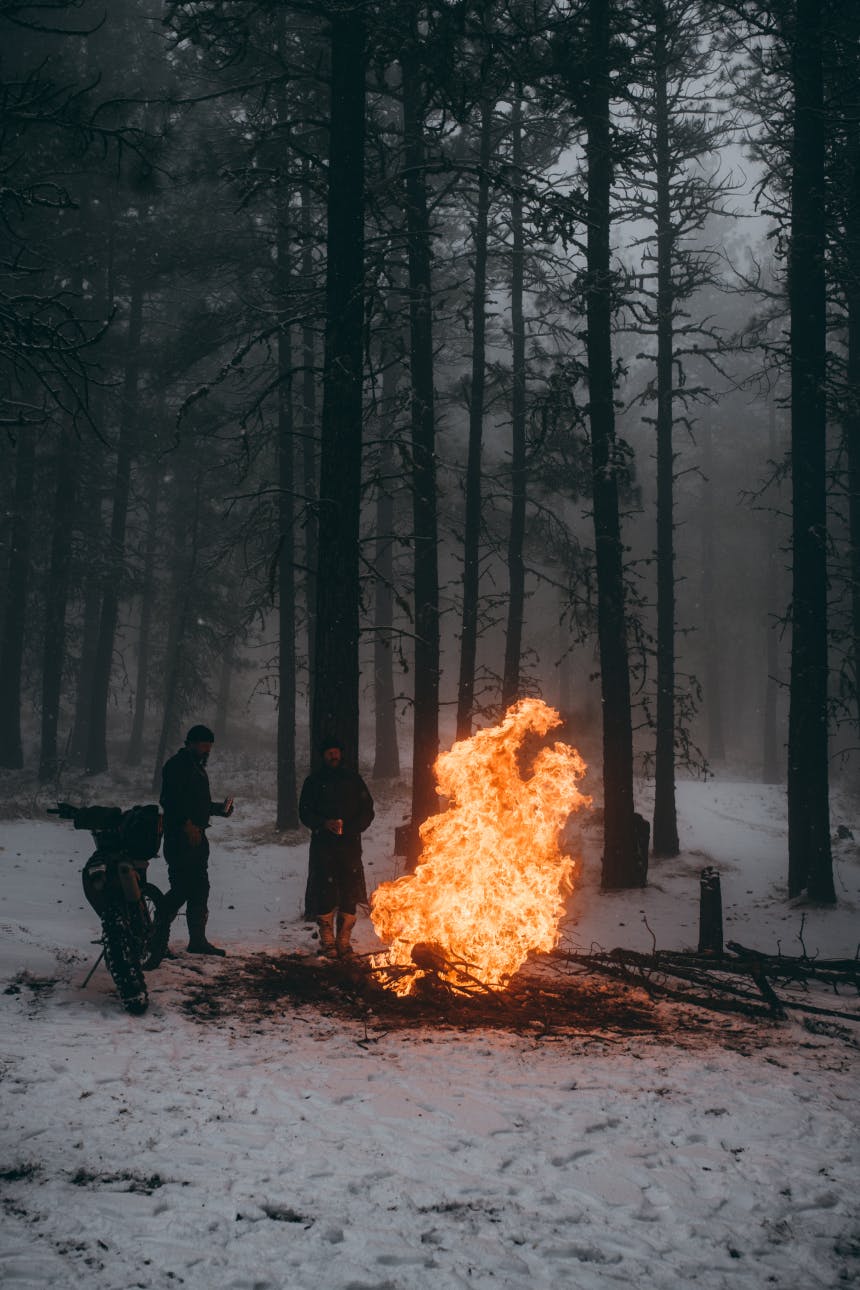 people standing by a motorcycle in a pine forest in a snow covered forest next to a large fire