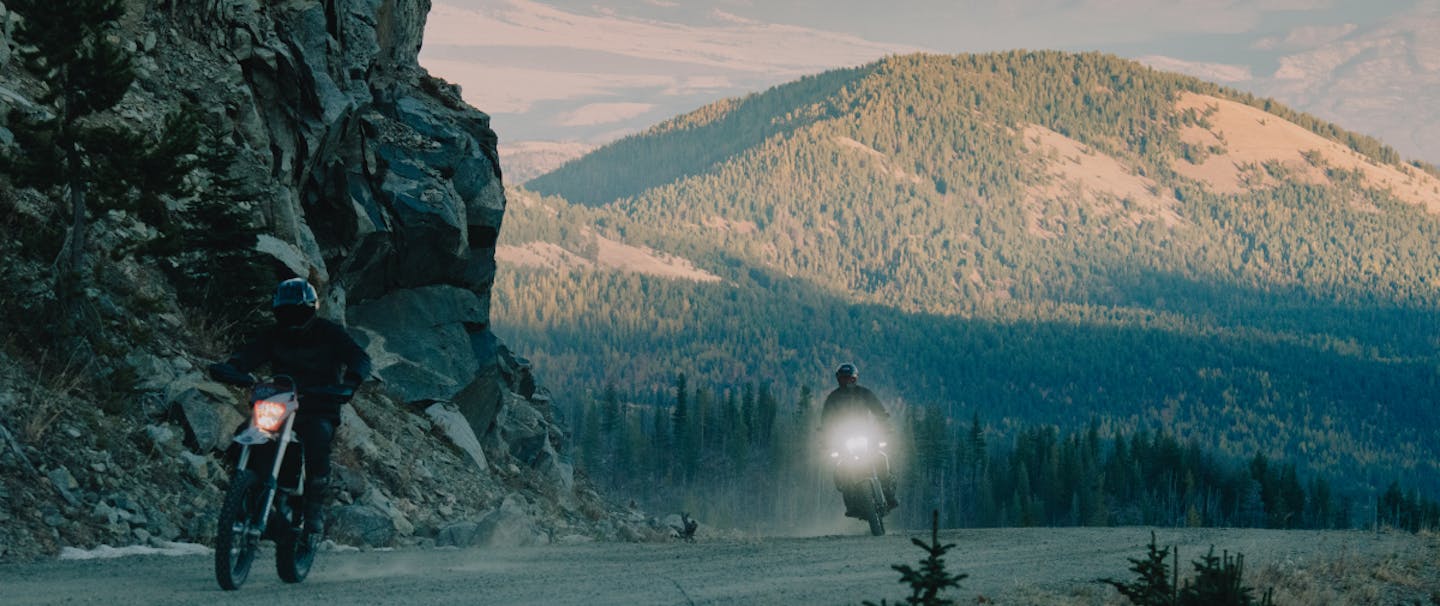 two people riding motorcycles on a dirt road with a pine covered hill in the background