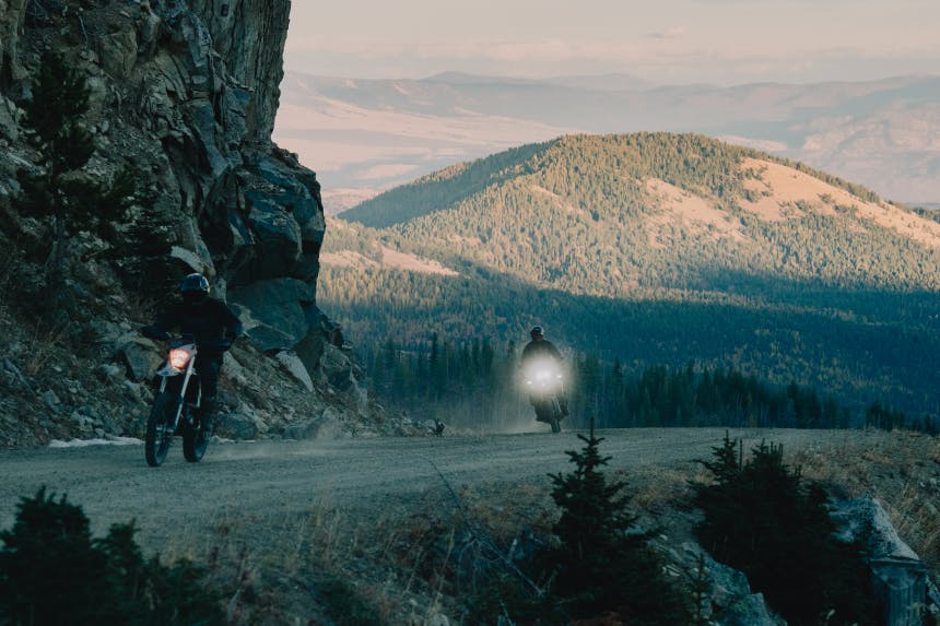 two people riding motorcycles on a dirt road with a pine covered hill in the background