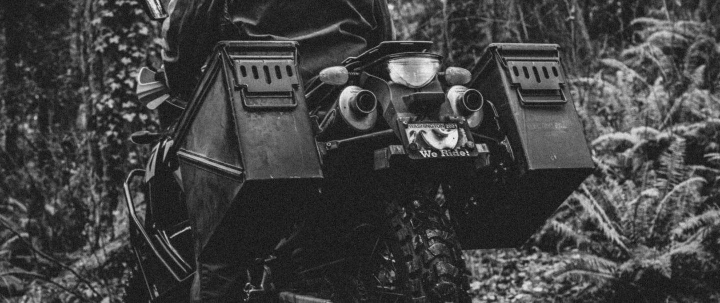 person sitting on motorcycle in the forest with ammunition cannisters mounted to the back wheel