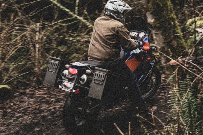 person in brwon jacket riding blue and orange motorcycle with ammo cannister mounted on back portion riding through forest