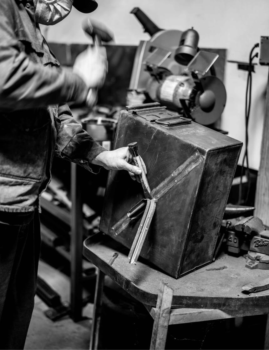 person in mask chiseling a metal detail off of the side of an ammo cannister in a workshop