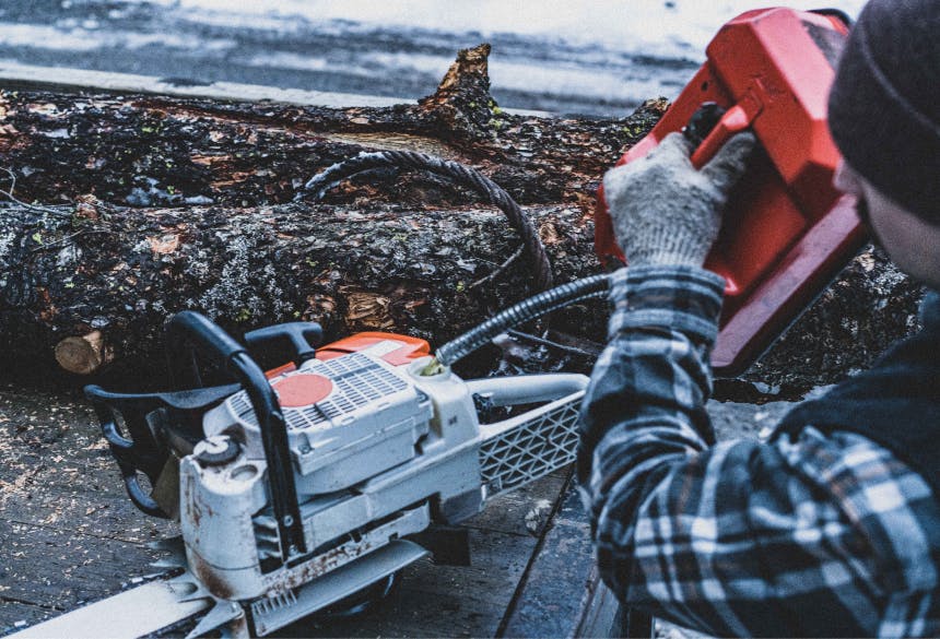 person using an orange tool on a chainsaw sitting on a table next to a downed log