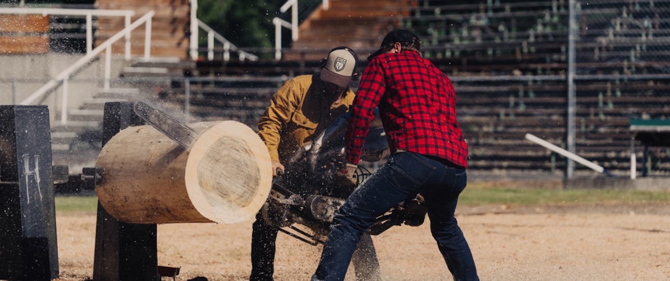 two men, one wearing a red and black plaid shirt with a black baseball cap and jeans, the other wearing a yellow Botton up, brown hat and jeans holding a souped up chainsaw cutting through a large log