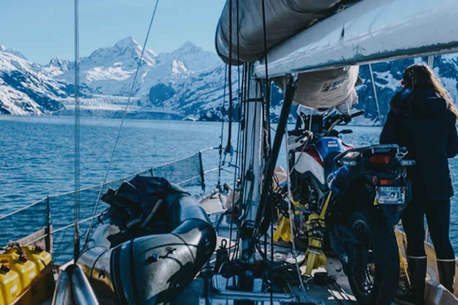 view from the deck of a working sailboat that has gear and a motorcycle strapped to the deck while a woman looks out to the snowy mountains