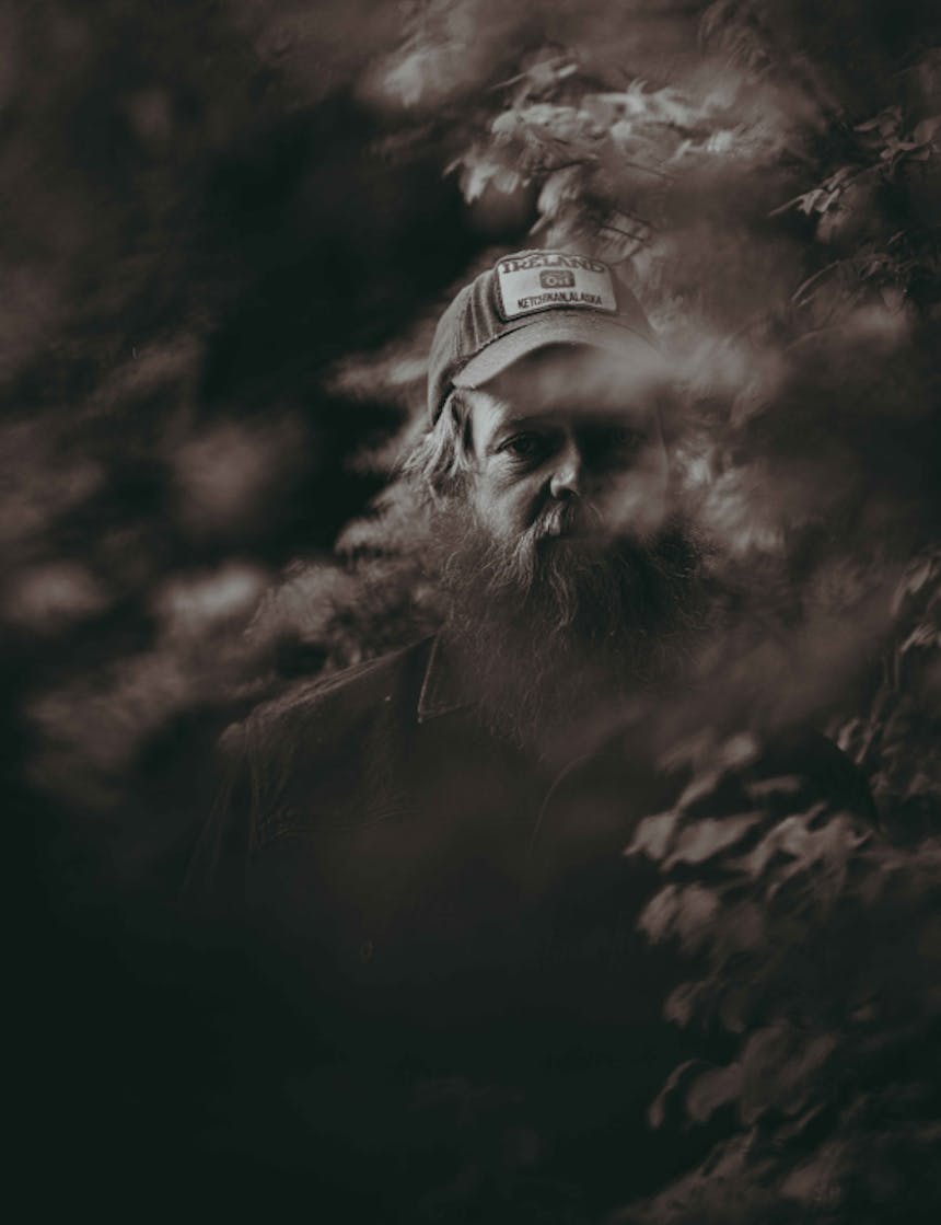 a black and white image of a bearded white man standing among the many leaves from branches on either side as he looks at the camera