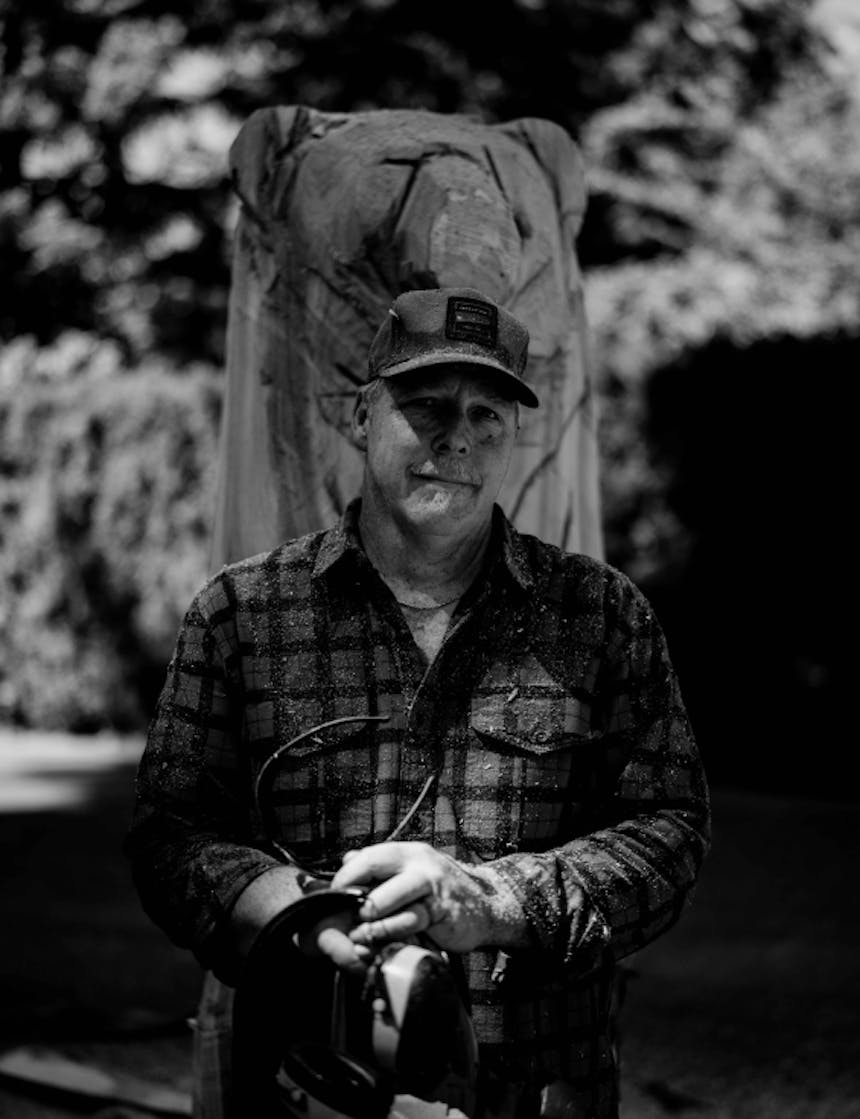 black and white of an older white man wearing a flannel shirt, holding his eye and ear protection standing in front of his wood carving of a bear in progress