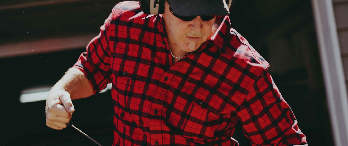 a close up of a white man wearing a black and red flannel shirt using a pull start for a chainsaw